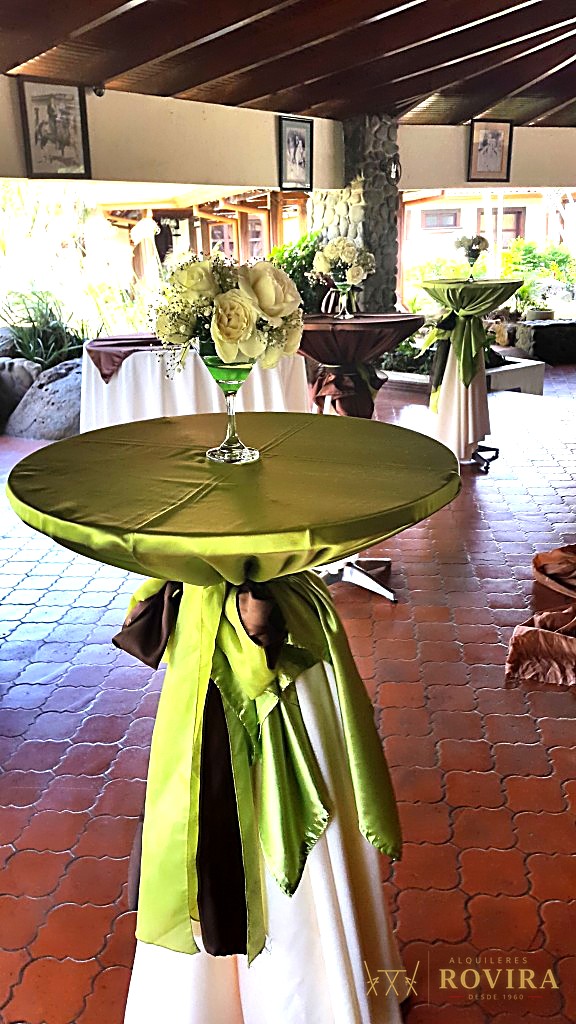 Mesa cocktelera alta y angosta, forrada en verde oliva con lazo café, centro floral blanco
