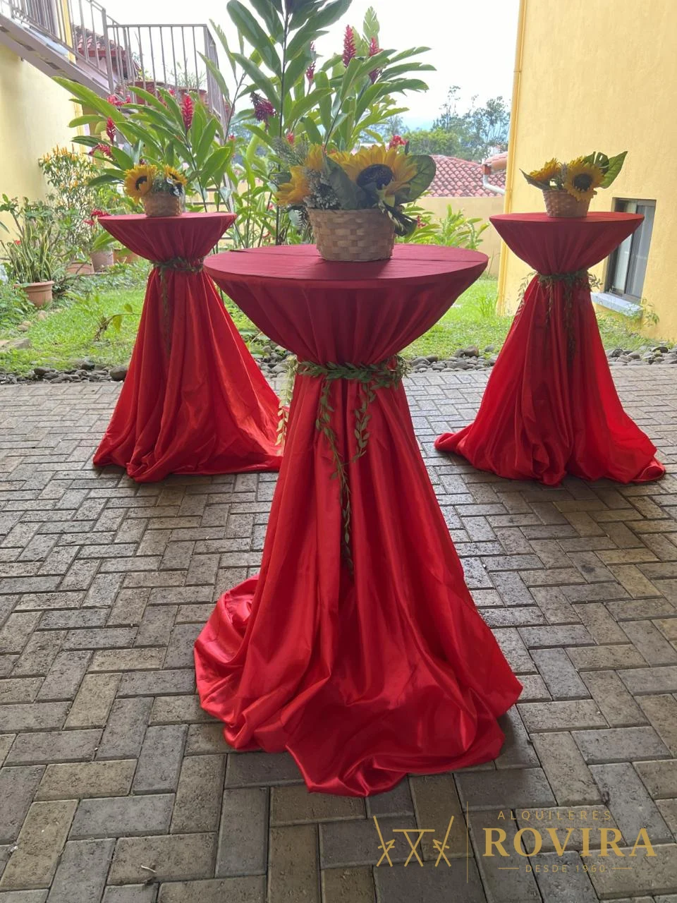Mesas cocktaileras altas con forro rojo y centros florales, evento en exterior