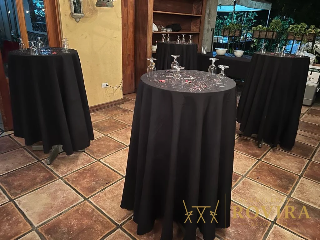 Mesas cocktaileras altas con forro negro y cristalería lista, montaje interior