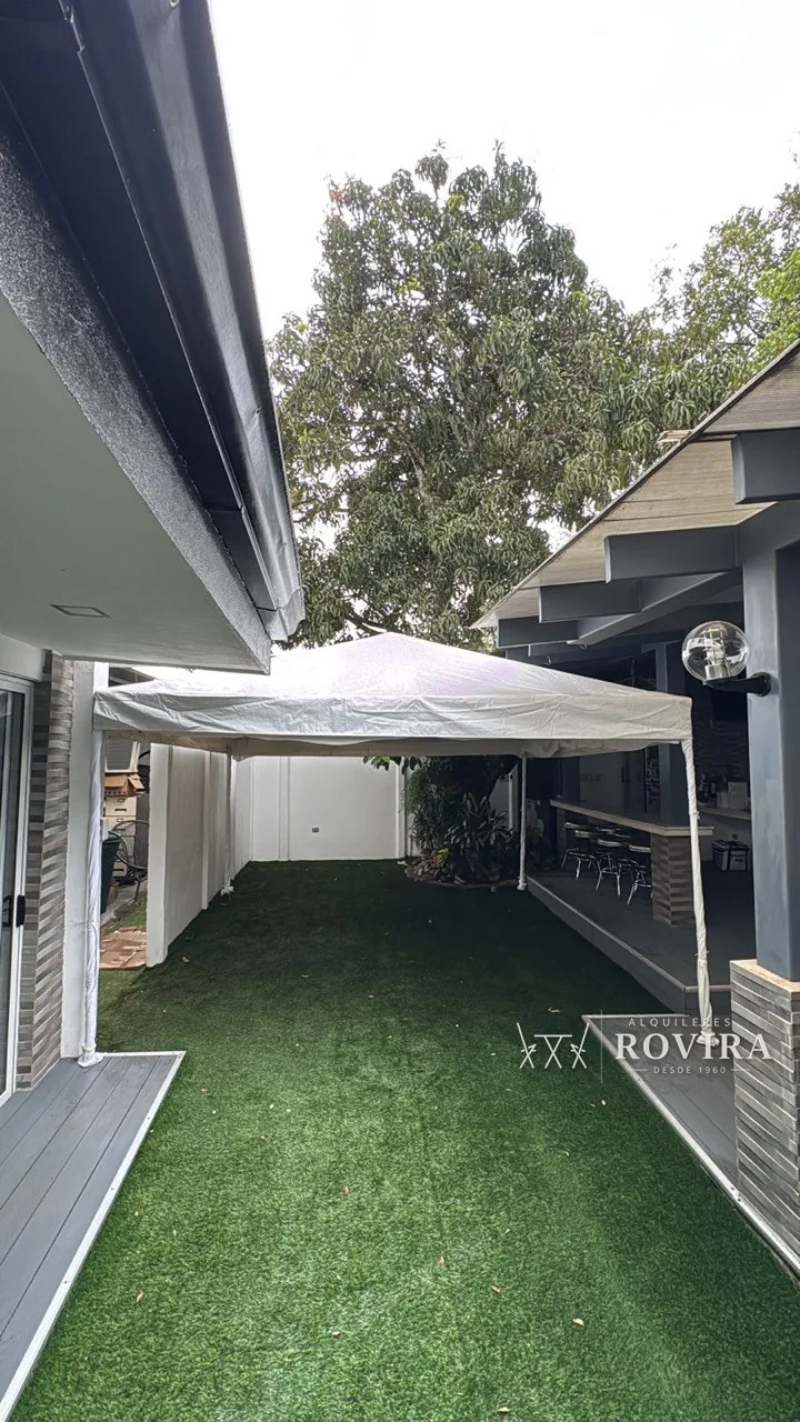Toldo blanco instalado en patio lateral techado con grama sintética, entre dos alas de una casa