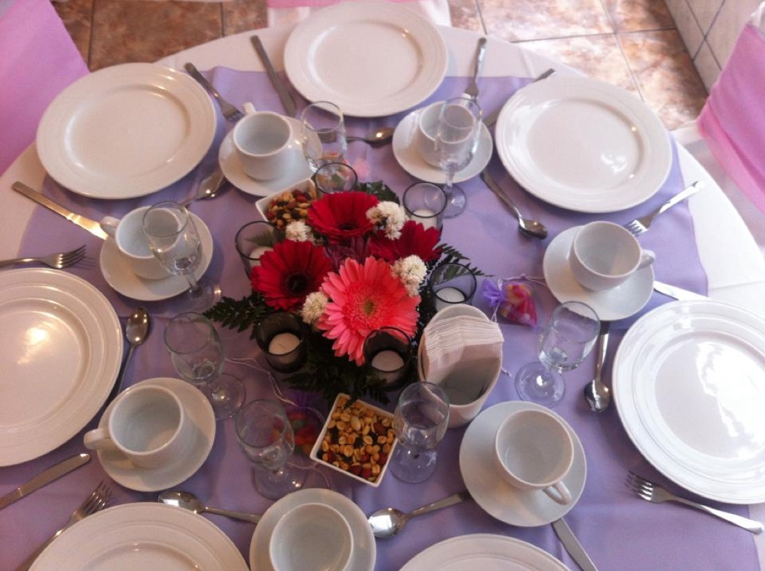 Mesa redonda vestida en lila con vajilla blanca, tazas de café y centro de flores rojas y rosadas