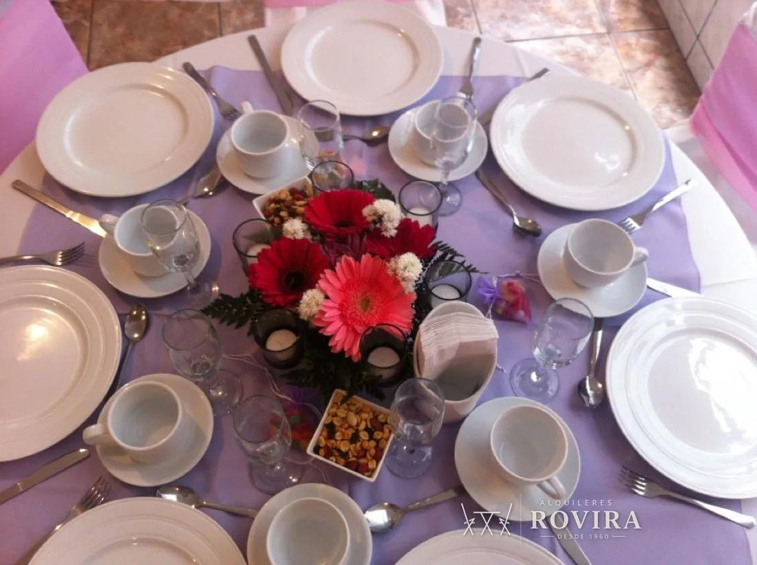 Mesa redonda vestida en lila con vajilla blanca, tazas de café y centro de flores rojas y rosadas