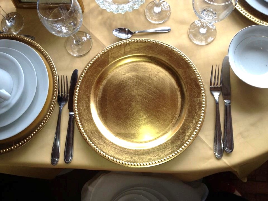 Primer plano de plato base dorado con vajilla blanca y copas de cristal, sobre mantel dorado