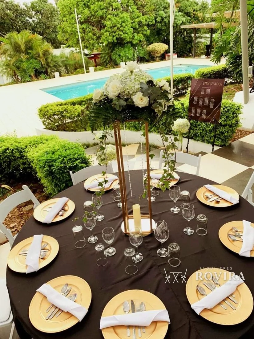 Mesa de boda al aire libre junto a piscina, platos base dorados, copas de vino y agua