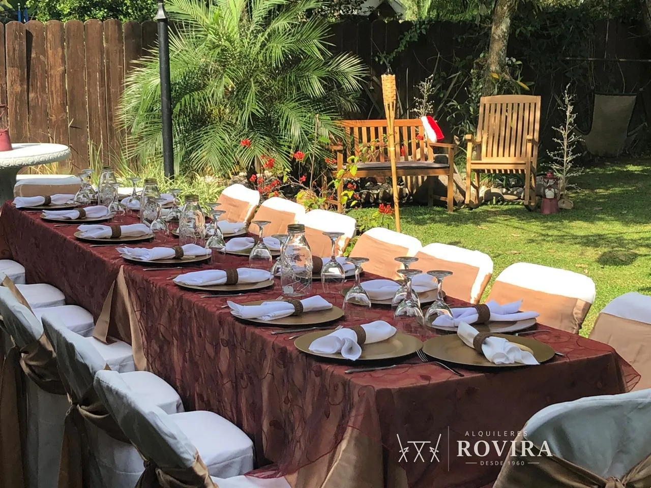 Mesa larga vestida en vino burgundy con platos dorados y jarras de vidrio, jardín con mobiliario de madera