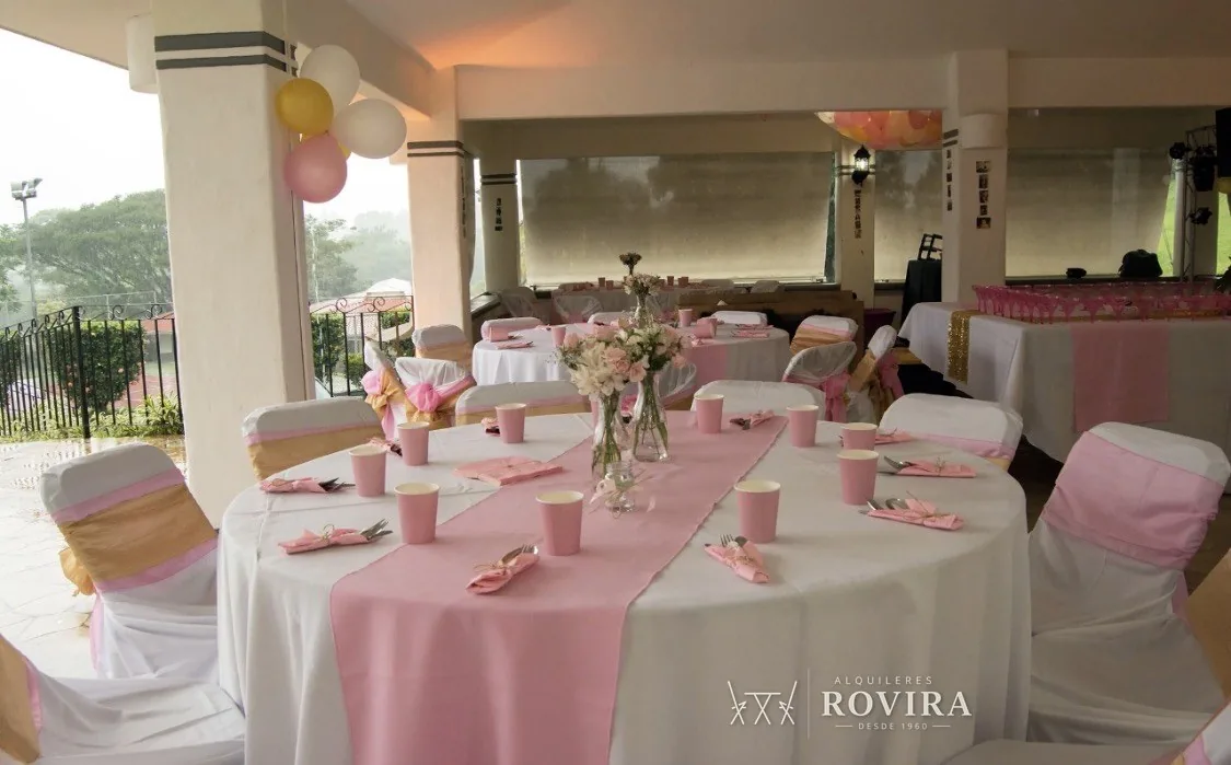 Mesas redondas vestidas de blanco con camino rosa, sillas con lazo rosa y dorado, terraza techada con vista al jardín y globos