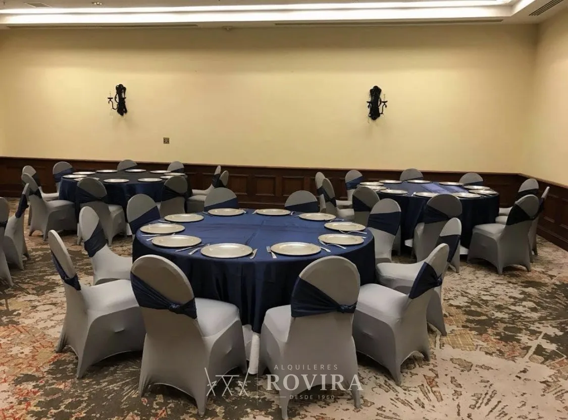 Salón formal tipo hotel con mesas redondas vestidas en azul marino, forros de silla gris con lazo azul marino y platos base, evento corporativo elegante