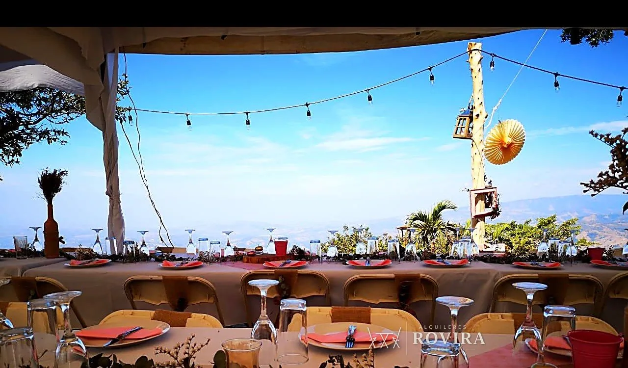 Mesa larga vestida con sillas cromadas de respaldar blanco y lazo café, sobre una montaña con vista panorámica
