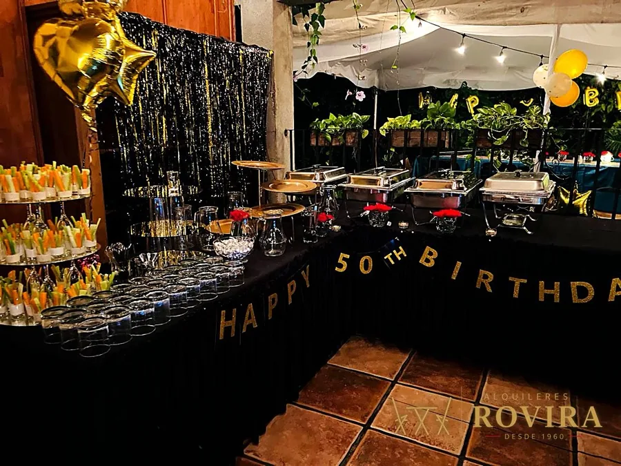 Buffet de cumpleaños 50 con chafing dishes, vegetales en vasitos y globos dorados y negros