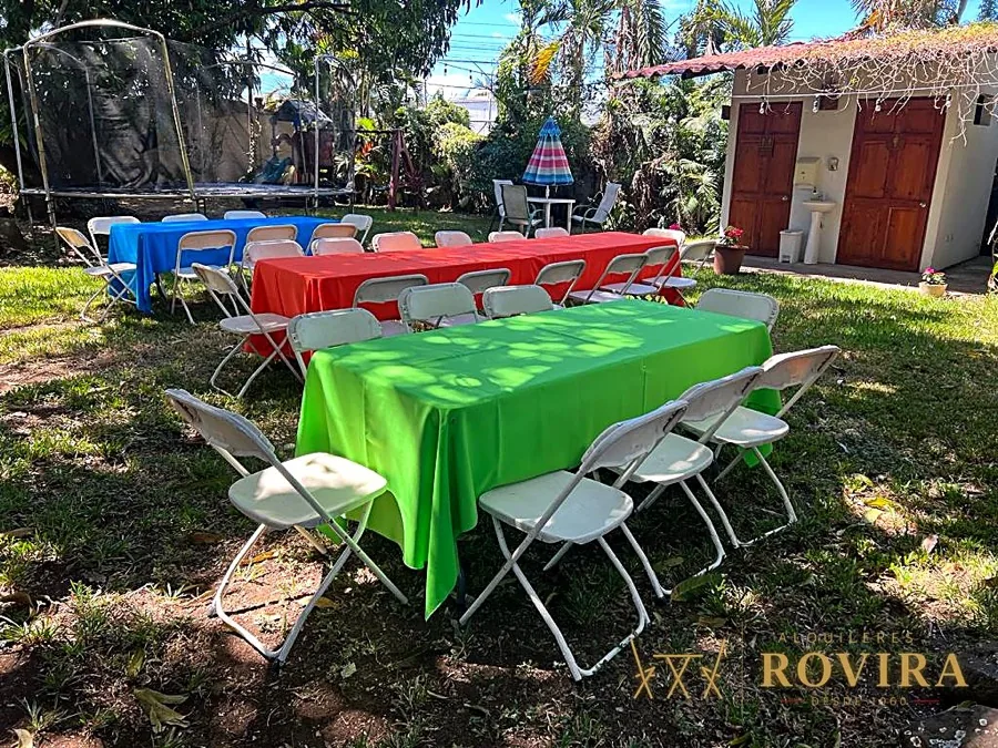 Mesas de colores (rojo, verde, azul) con sillas blancas en jardín